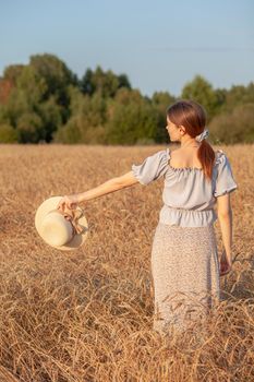 Young beautiful girl with long curly hair poses in a wheat field in the summer at sunset. A girl holds a hat in her hand against the background of a wheat field. The view from the back. Toning.