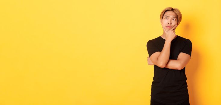 Portrait of thoughtful attractive guy in black t-shirt looking upper left corner. Young man pondering idea, standing yellow background.