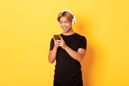 Young smiling asian guy using smartphone and listening music or podcast in wireless headphones, standing yellow background.
