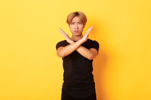 Portrait of disappointed frowning asian man, making cross gesture to stop something bad, standing yellow background.