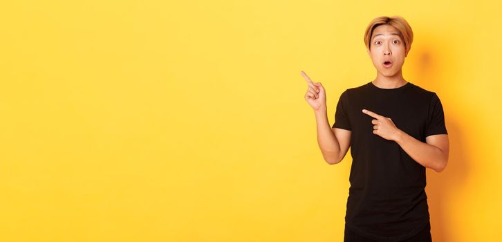 Portrait of impressed and excited handsome asian guy in black t-shirt, react to your logo, pointing fingers upper left corner.