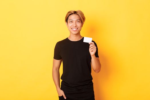 Cheerful handsome asian guy smiling pleased and showing credit card, standing yellow background.
