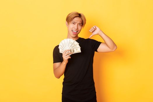 Sassy handsome asian guy pointing finger at cash and looking pleased. Korean man borrowed money, standing yellow background.