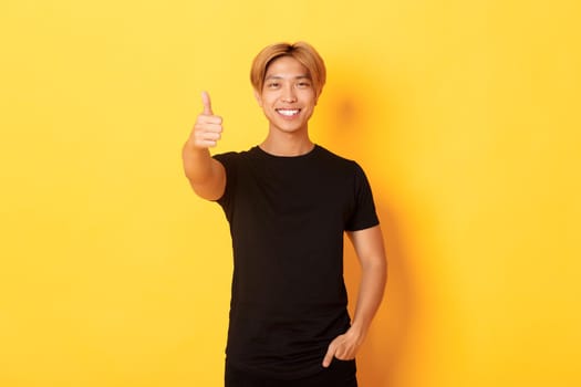Portrait of satisfied asian man with blond hair, standing over yellow background and showing thumbs-up.