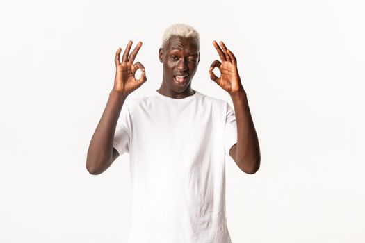 Portrait of sassy attractive african-american blond man, winking and showing okay gesture in approval, standing white background.