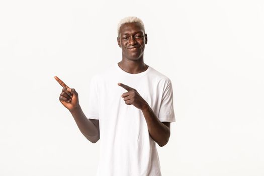 Portrait of young african american blond guy with smug face, smirking and pointing upper left corner pleased, standing white background.