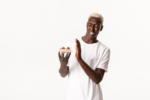 Portrait of disgusted handsome african american young blond man, unwilling eat dessert, cringe from disgusting cake, standing white background.