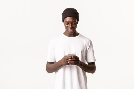 Portrait of handsome african-american young man in beanie using mobile phone, messaging and smiling at smartphone screen, white background.