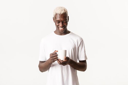 Portrait of relaxed happy african-american blond guy, looking dreamy and smiling at cup of coffee, standing white background.