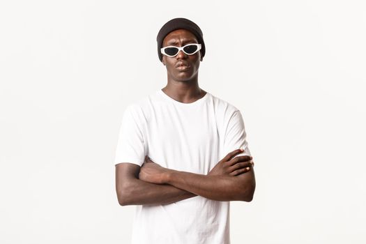 Portrait of skeptical, serious-looking Black guy in sunglasses and beanie, standing with arms crossed and frowning doubtful, express disbelief, white background.