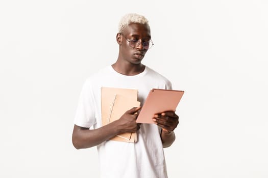 Portrait of determined, serious-looking african american male student, looking focused at digital tablet, studying, holding notebooks, standing white background.
