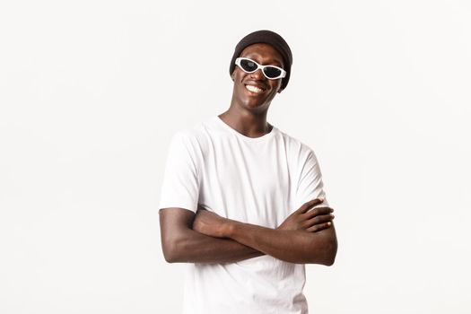 Portrait of sassy and stylish african-american guy in beanie, sunglasses, standing pleased with arms crossed and smiling happy at camera, white background.