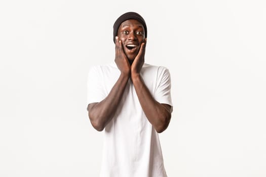 Portrait of happy surprised young african-american guy, looking amazed and cheerful, gasping fascinated, holding hands on cheeks, white background.
