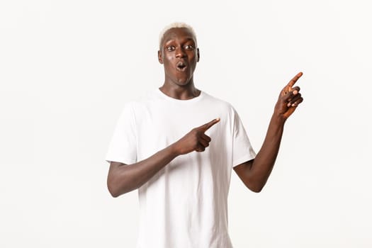 Portrait of surprised and amazed african-american blond man, looking impressed and pointing fingers upper right corner, white background.