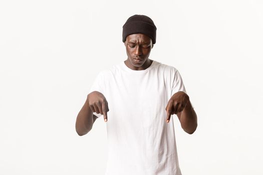 Portrait of confused african-american guy in beanie, looking perplexed and pointing fingers down, standing puzzled over white background.