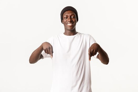 Portrait of cheerful attractive Black young man in beanie, pointing fingers down and smiling happy, showing logo, white background.