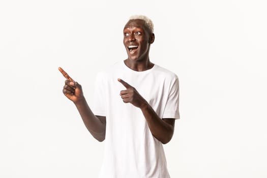 Portrait of excited handsome african-american blond guy, looking happy and surprised at logo, pointing fingers upper left corner, white background.