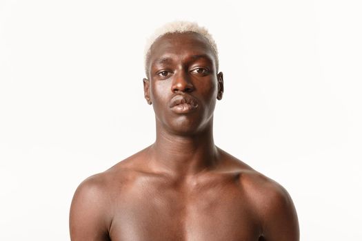Close-up of serious-looking handsome african-american guy with blond hair, standing with bare chest over white background, looking determined.