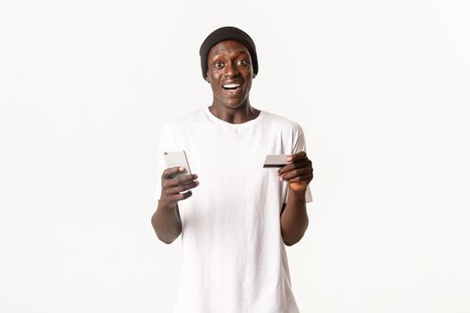 Portrait of excited, happy african-american young male student, holding credit card and smartphone with amused smile, white background.