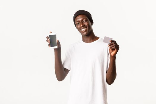 Portrait of pleased, smiling african-american guy in beanie, showing smartphone screen and credit card, white background.