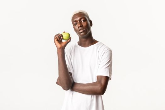 Portrait of handsome sassy african-american young blond man, holding green apple and looking at camera, eating healthy, standing white background.