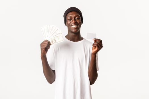 Portrait of happy, lucky african-american male in beanie, smiling cheerful while showing credit card with money, white background.
