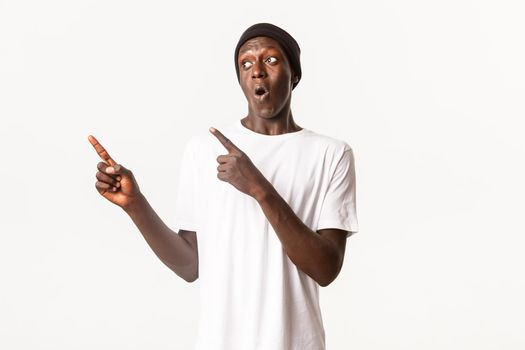 Portrait of excited and pleased african-american guy in beanie, pointing fingers and looking left with intrigued expression, white background.