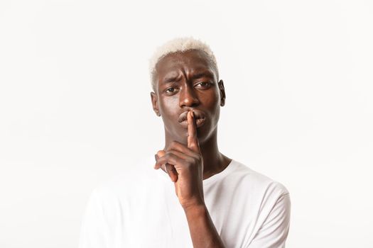 Portrait of worried african-american blond guy making warning, shushing at person and frowning displeased, hushing, quiet please, standing white background.