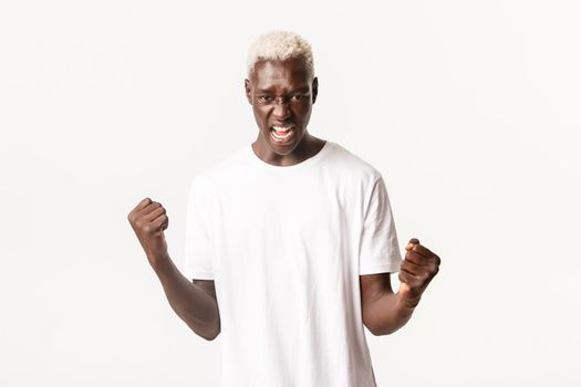 Portrait of confident and strong african-american blond man, clenching fists and shouting encouraged, standing white background.