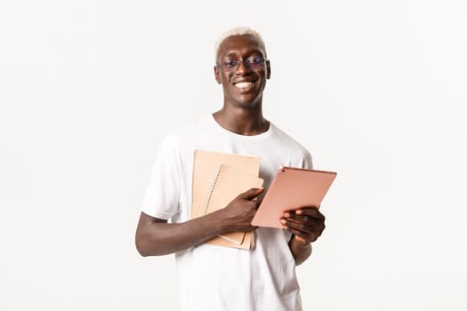 Portrait of handsome smiling young african-american blond guy, wearing glasses. Student holding notebooks and digital tablet, looking cheerful at camera, white background.