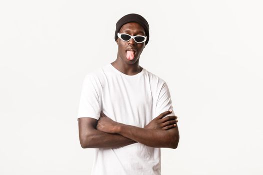 Portrait of funny and cool african-american guy in beanie and sunglasses, showing tongue with unbothered expression, standing white background.