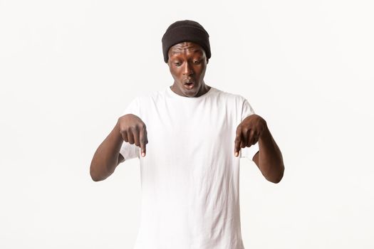 Portrait of wondered and amazed african-american guy in beanie, handsome man pointing fingers down and looking surprised, standing white background.