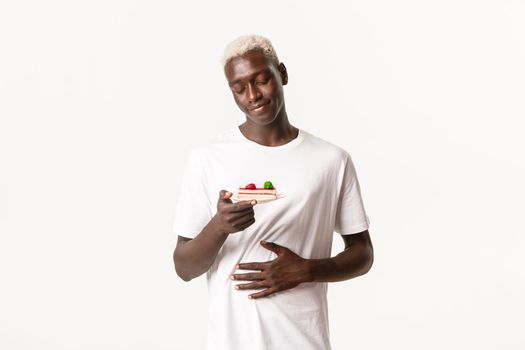 Portrait of delighted and happy african-american blond guy foodie, rubbing tummy and smiling at delicious cake, standing white background.