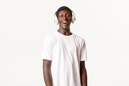Portrait of amused and happy african-american young guy listening podcast or music in headphones, smiling pleased, white background.