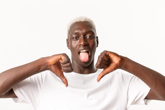 Close-up of disappointed african-american blond male showing thumbs-down and stick tongue from disappointment, dislike and judging something bad, white background.