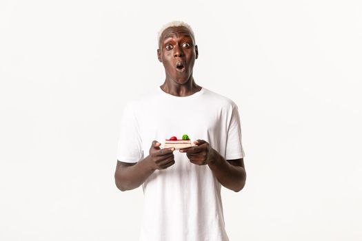 Portrait of surprised african-american blond guy, say wow, looking wondered and holding piece of cake, standing white background.