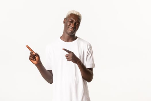 Portrait of skeptical and unamused african-american blond guy, tilt head and grimacing bothered, pointing fingers upper left corner, standing white background.