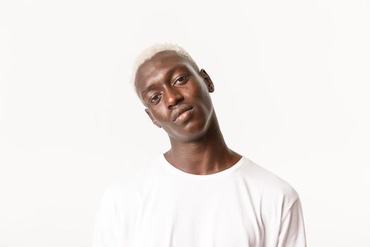 Close-up of reluctant african-american blond man, tilt head and looking at camera skeptical, standing unamused over white background.
