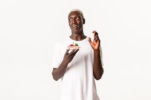 Portrait of hopeful attractive Black guy celebrating birthday, making wish and holding b-day cake, cross fingers for dream come true, white background.