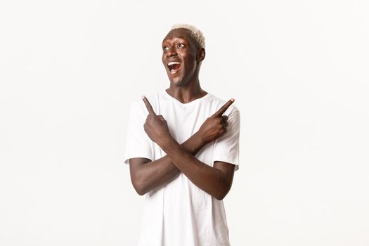 Portrait of excited and amused african-american blond guy, looking left amazed and smiling, pointing fingers sideways, showing variants, white background.