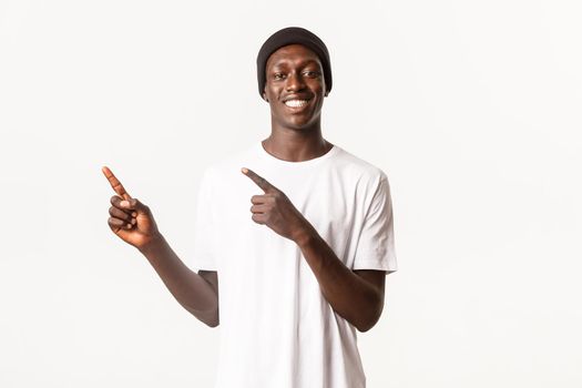 Portrait of cheerful handsome african-american male in beanie, pointing fingers upper left corner and smiling, showing logo or advertisement, white background.