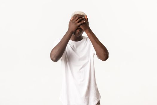 Portrait of gloomy and sad african-american blond guy, shut eyes with hands and looking miserable, standing white background.