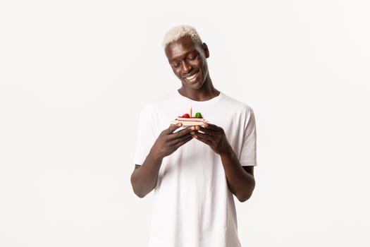 Portrait of cute and pleased smiling african-american blond man, celebrating birthday, looking happy at b-day cake, standing white background and making wish.