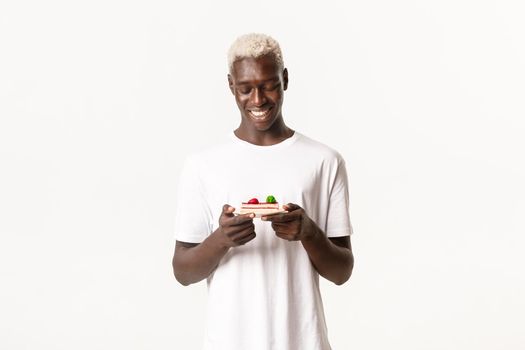 Portrait of attractive blond african-american guy, smiling happy and looking at delicious piece of cake, standing white background.