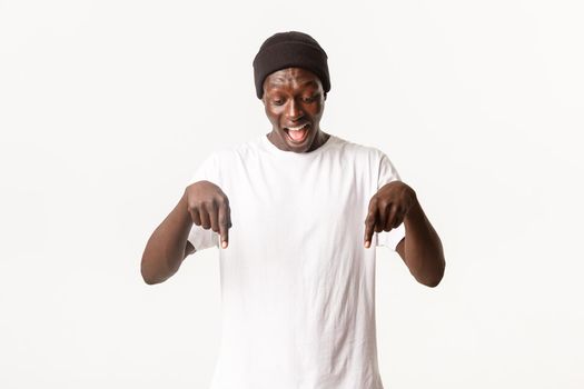 Portrait of excited, happy african-american man in beanie, yelling from amazement and pointing fingers down, looking at your logo cheerful, standing white background.