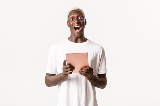 Portrait of excited handsome african-american male student in glasses, looking amazed, holding digital tablet, standing white background.