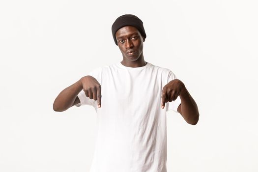 Portrait of serious african-american guy in beanie, looking at camera and pointing fingers down with poker face, white background.