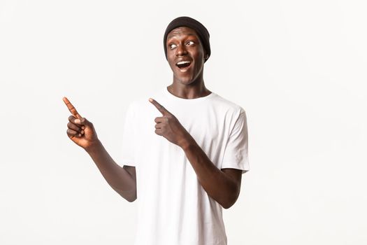 Portrait of excited and pleased african-american guy in beanie, pointing fingers and looking left with intrigued expression, white background.