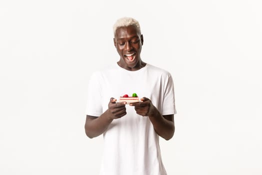 Portrait of excited, happy african-american male with blond hair, looking amazed at delicious cake, smiling cheerful, standing white background.