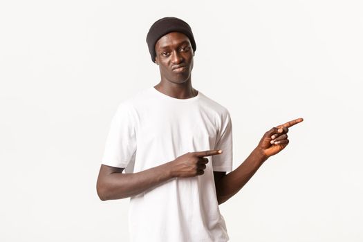 Portrait of skeptical and judgemental african-american guy in beanie, pointing fingers right and grimacing disappointed, express dislike, standing white background.